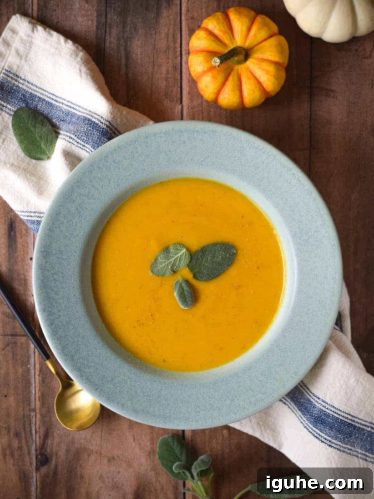 Golden Harvest Squash Soup 11 Overhead view of a blue bowl of creamy squash soup, garnished with sage, on a wooden background with a spoon.