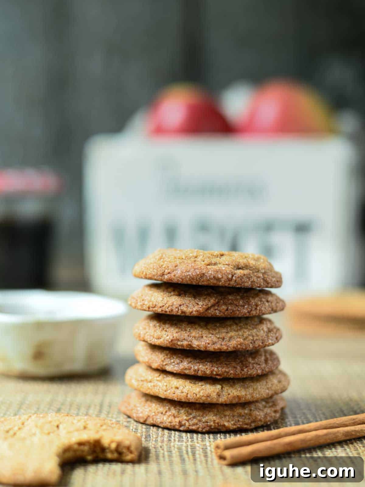 A tall stack of golden-brown apple cider cookies, with a rustic bucket of fresh apples in the softly blurred background, evoking a cozy autumn scene.