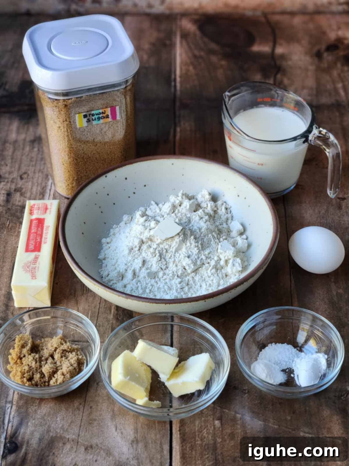 Decadent Brown Sugar Stacks 3 Ingredients for brown sugar pancakes laid out on a wooden surface, including flour, brown sugar, buttermilk, eggs, and butter.