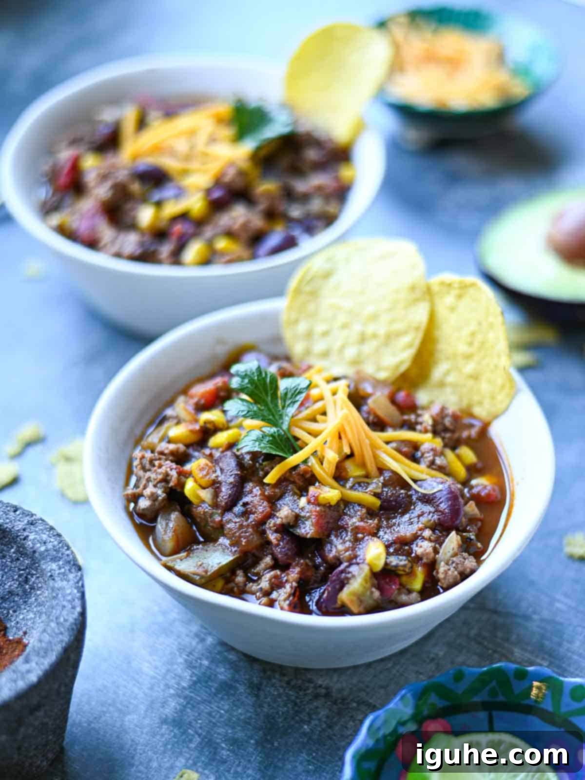Fire-roasted chili in white bowls on a blue surface, garnished with fresh herbs and a lime wedge.