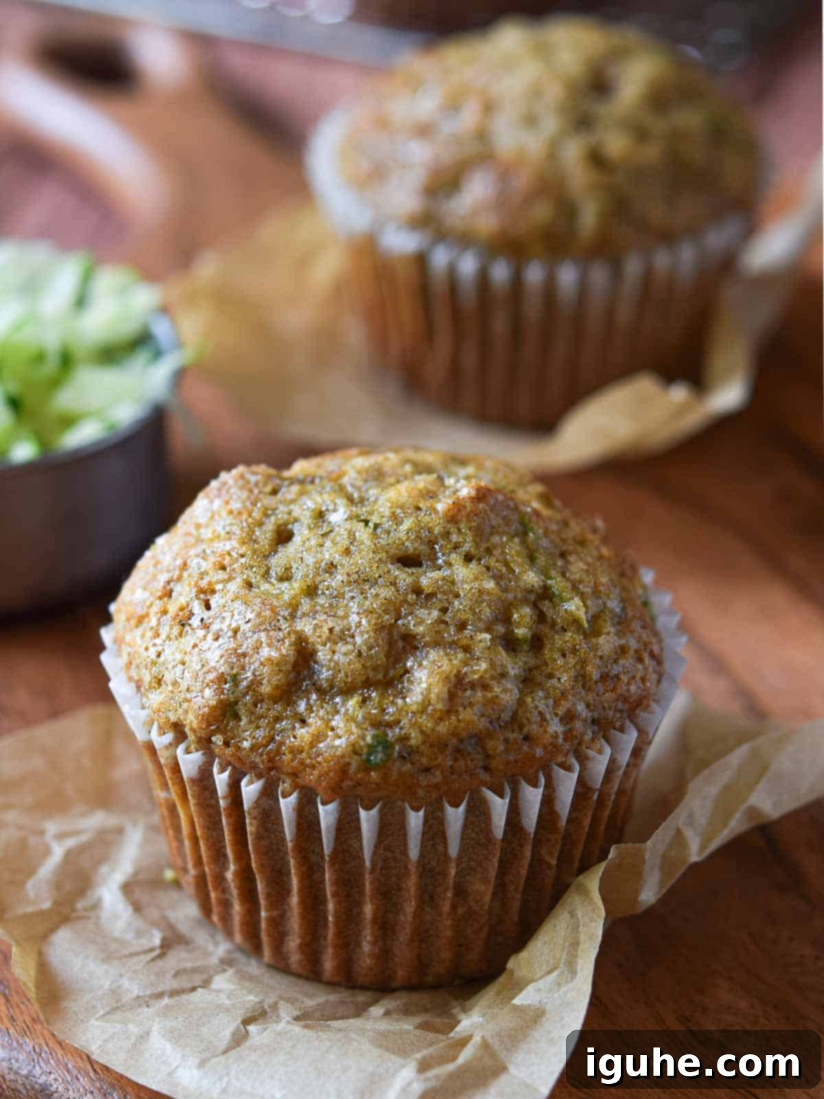 Apple-Kissed Zucchini Muffins 11 Two muffins on a wood surface with shredded zucchini in the background.