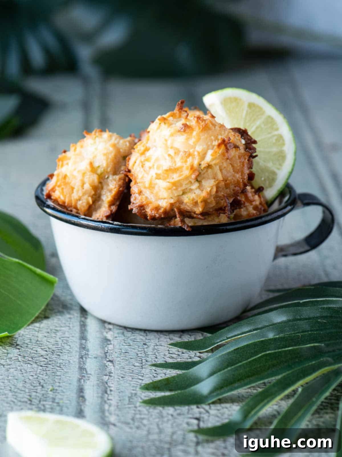Tropical Coconut Lime Macaroons 11 Coconut macaroons in a white cup with palm leaves and limes next to it, creating a tropical dessert scene.