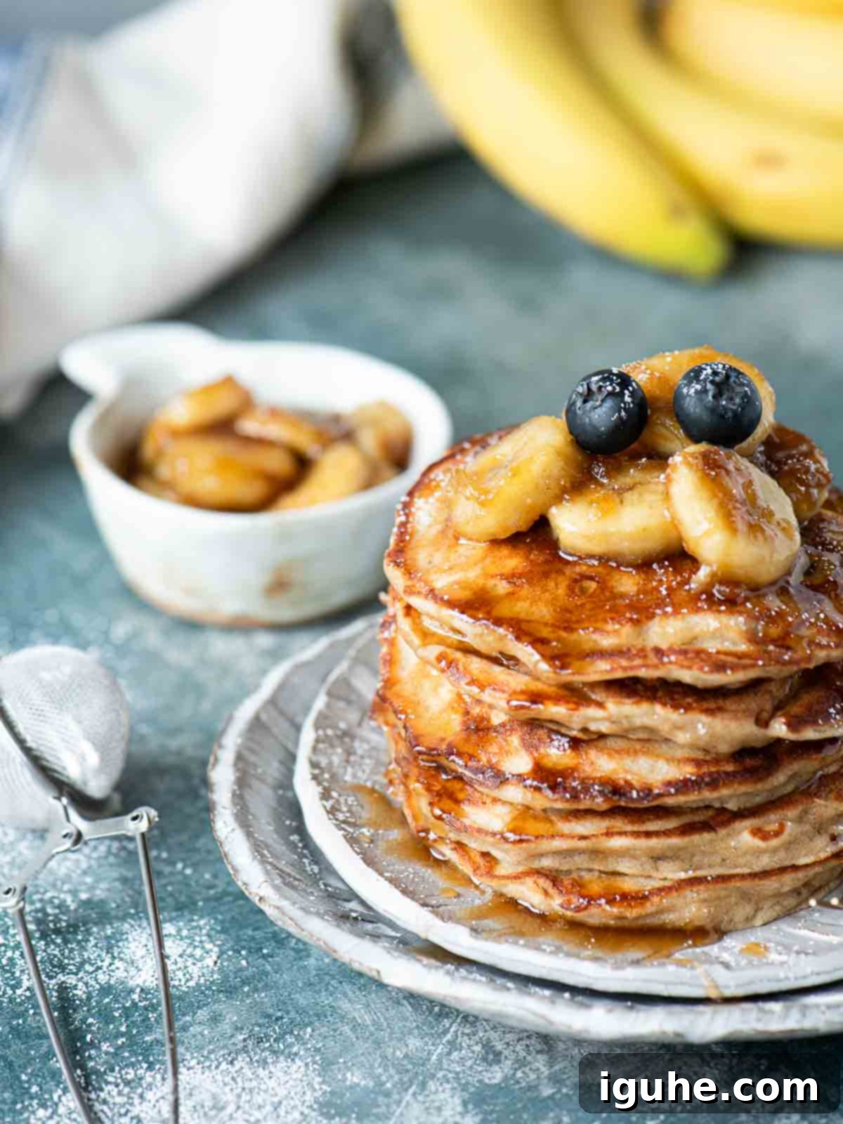 A stack of golden brown banana bread pancakes, generously topped with glistening caramelized banana compote and a few fresh banana slices, served on a modern grey plate against a blue backdrop, ready for a delicious breakfast or brunch.