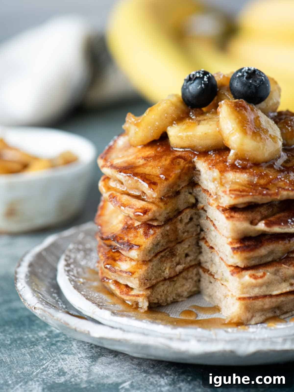 Homemade Banana Bread Pancakes with Banana Compote 7 A beautifully presented stack of banana bread pancakes, cut open to reveal the fluffy interior, generously topped with banana slices and a light drizzle of syrup. Served on a white plate with a fork.