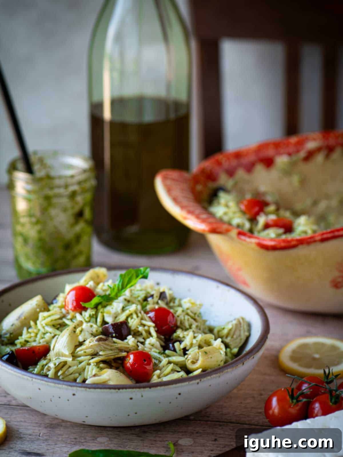 A vibrant bowl of Orzo Pesto Salad, featuring rice-shaped pasta, cherry tomatoes, feta, artichokes, and olives, dressed in green pesto, placed on a rustic kitchen table.