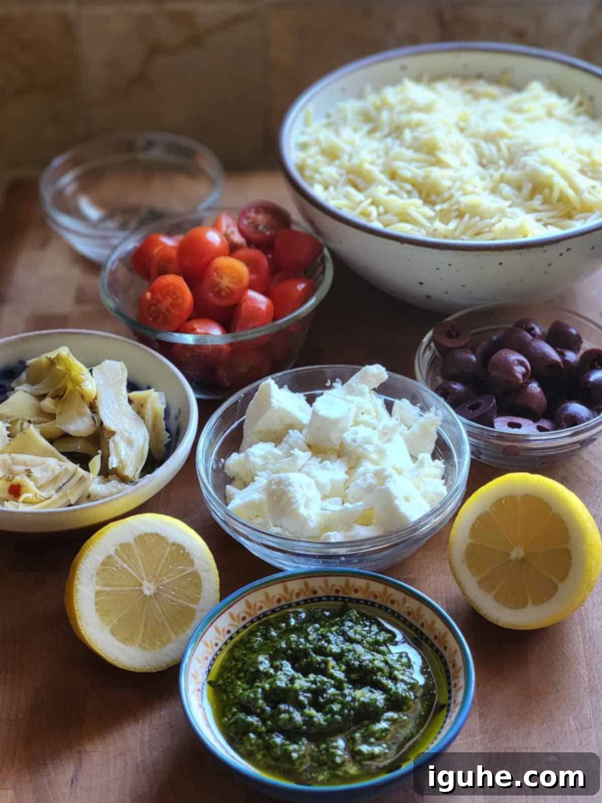 Vibrant Pesto Orzo Bowl 3 Assortment of fresh ingredients for Orzo Pesto Salad, including dry orzo pasta, cherry tomatoes, feta, artichokes, and olives, laid out on a rustic wooden board.