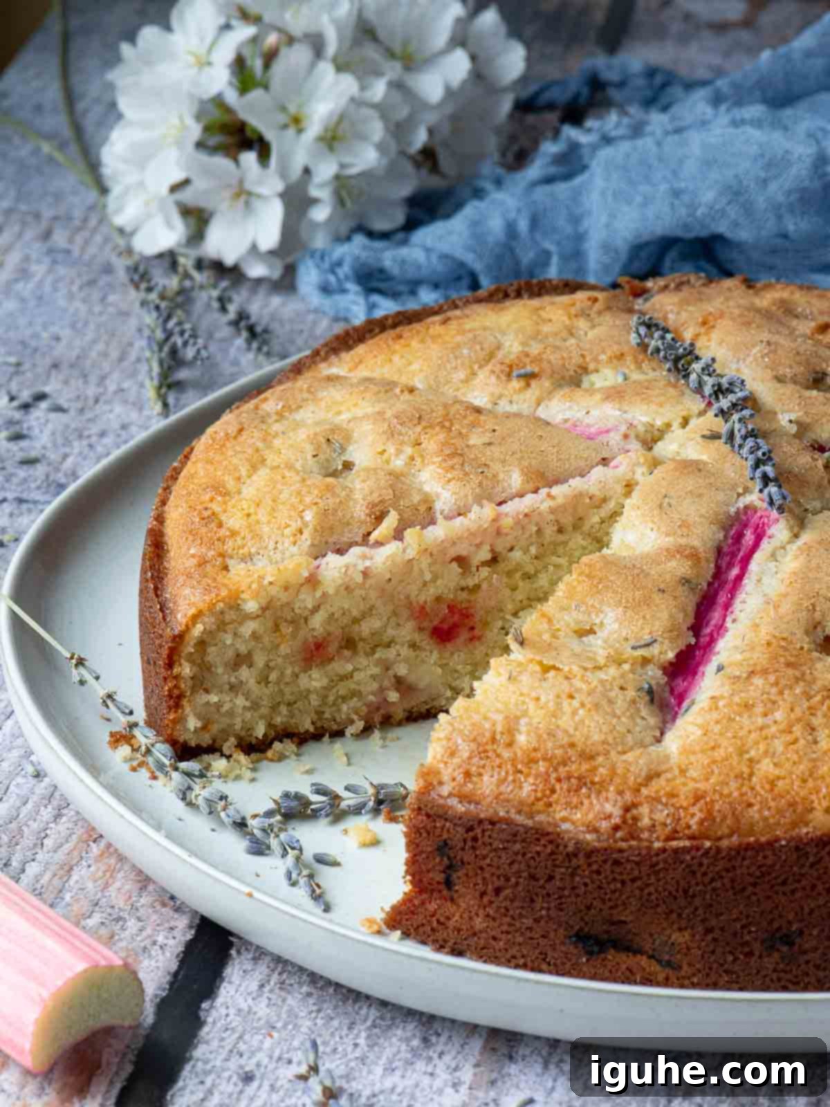 Rhubarb and Lavender Cake 7 Rhubarb lavender cake with a slice removed on a wood surface with flowers in the back.