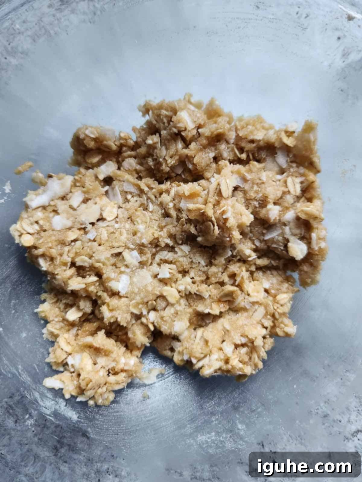 Close-up of thick, sticky batter for coconut oatmeal cookies in a clear mixing bowl, showing flecks of oats and coconut.