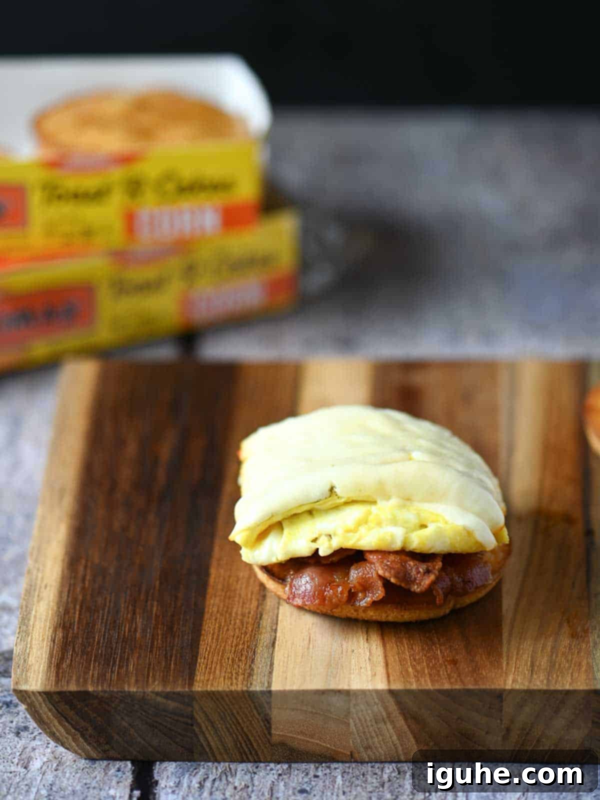 A stack of ingredients for the breakfast sandwich: a perfectly folded egg and a slice of melting cheese, resting on top of crispy bacon and a corn toaster cake.