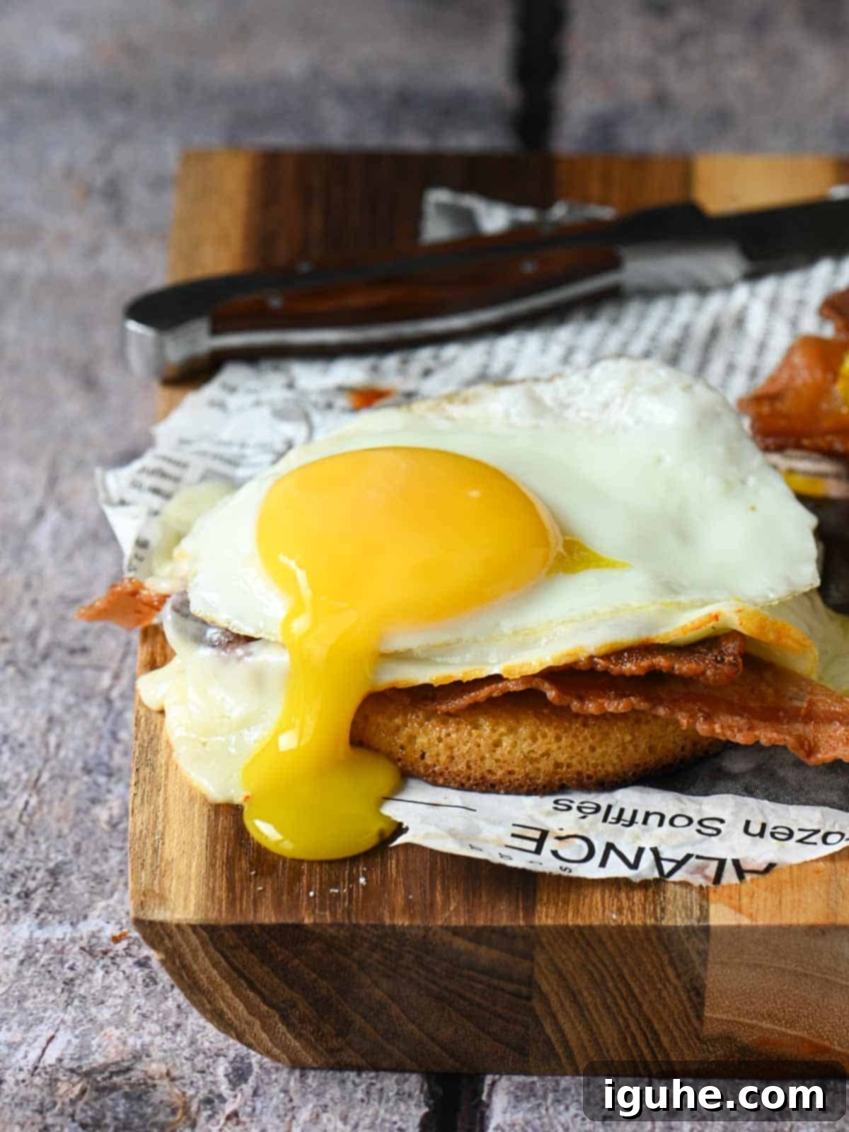 An open-faced serving of a toaster cake breakfast, featuring a beautifully soft-boiled egg with a visibly runny yolk, drizzled with maple syrup, alongside crispy bacon and melted cheese, presented on a wooden cutting board.