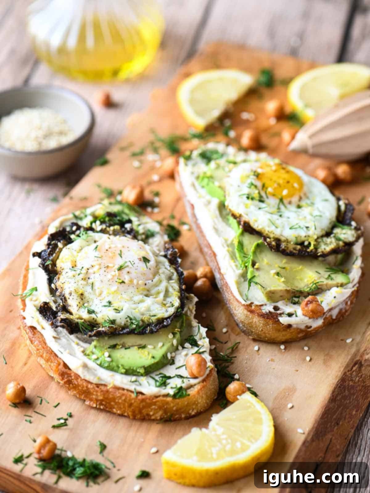 Two slices of gourmet hummus toast artfully arranged on a wooden cutting board, showcasing the vibrant colors of za'atar fried eggs, creamy avocado, lemon slices, and fresh dill.