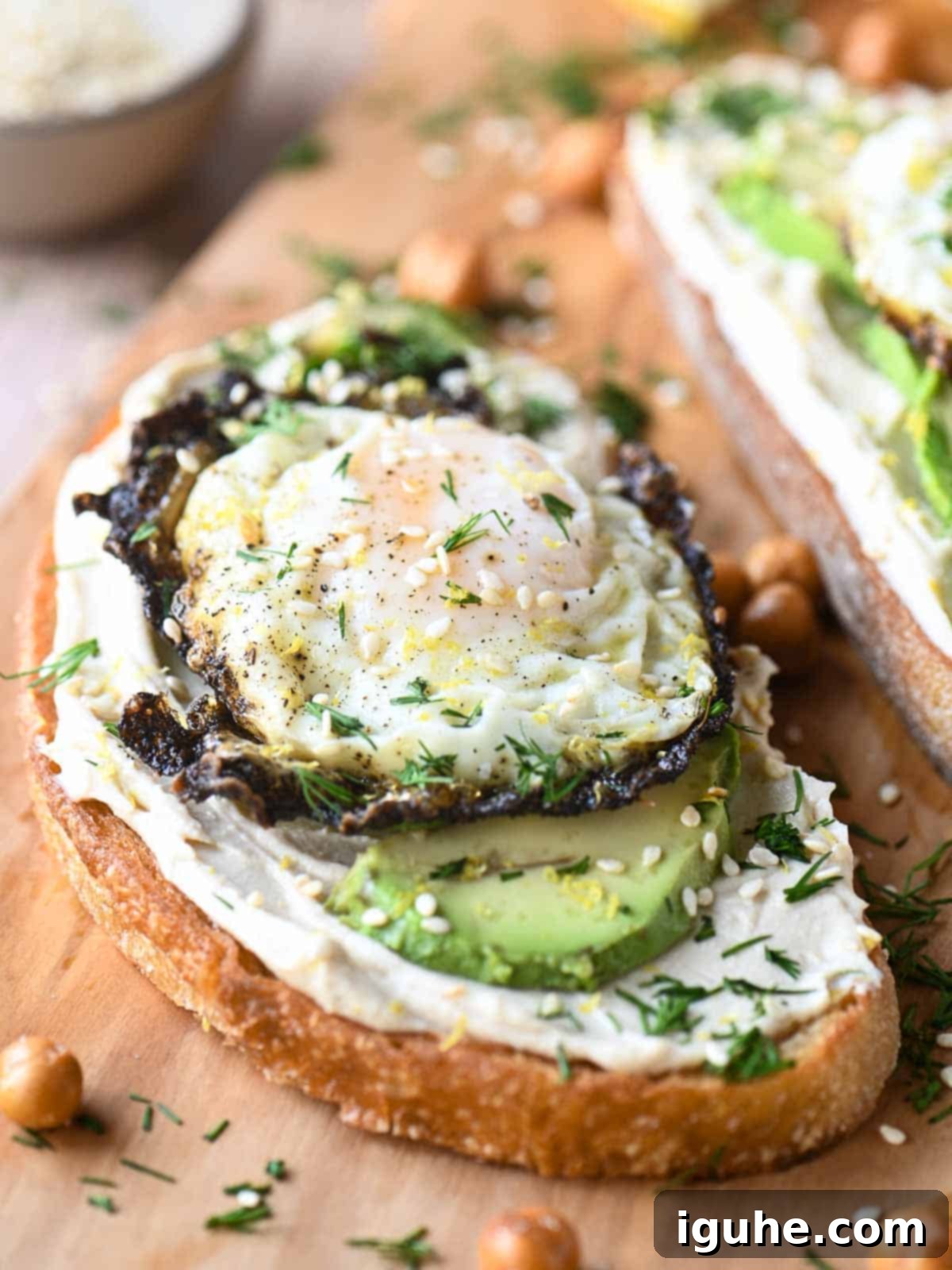 A close-up shot of hummus toast on a wooden cutting board, showcasing the textures of the creamy hummus, perfectly fried egg, smooth avocado, and crunchy dried chickpeas. The chickpeas add visual interest and a delightful nutty flavor.