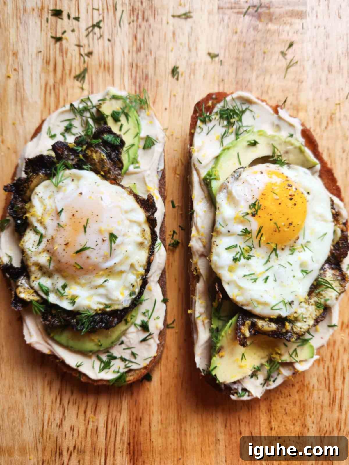 Two slices of perfectly assembled hummus toast with za'atar fried eggs, creamy avocado, and a final flourish of fresh dill and sesame seeds.