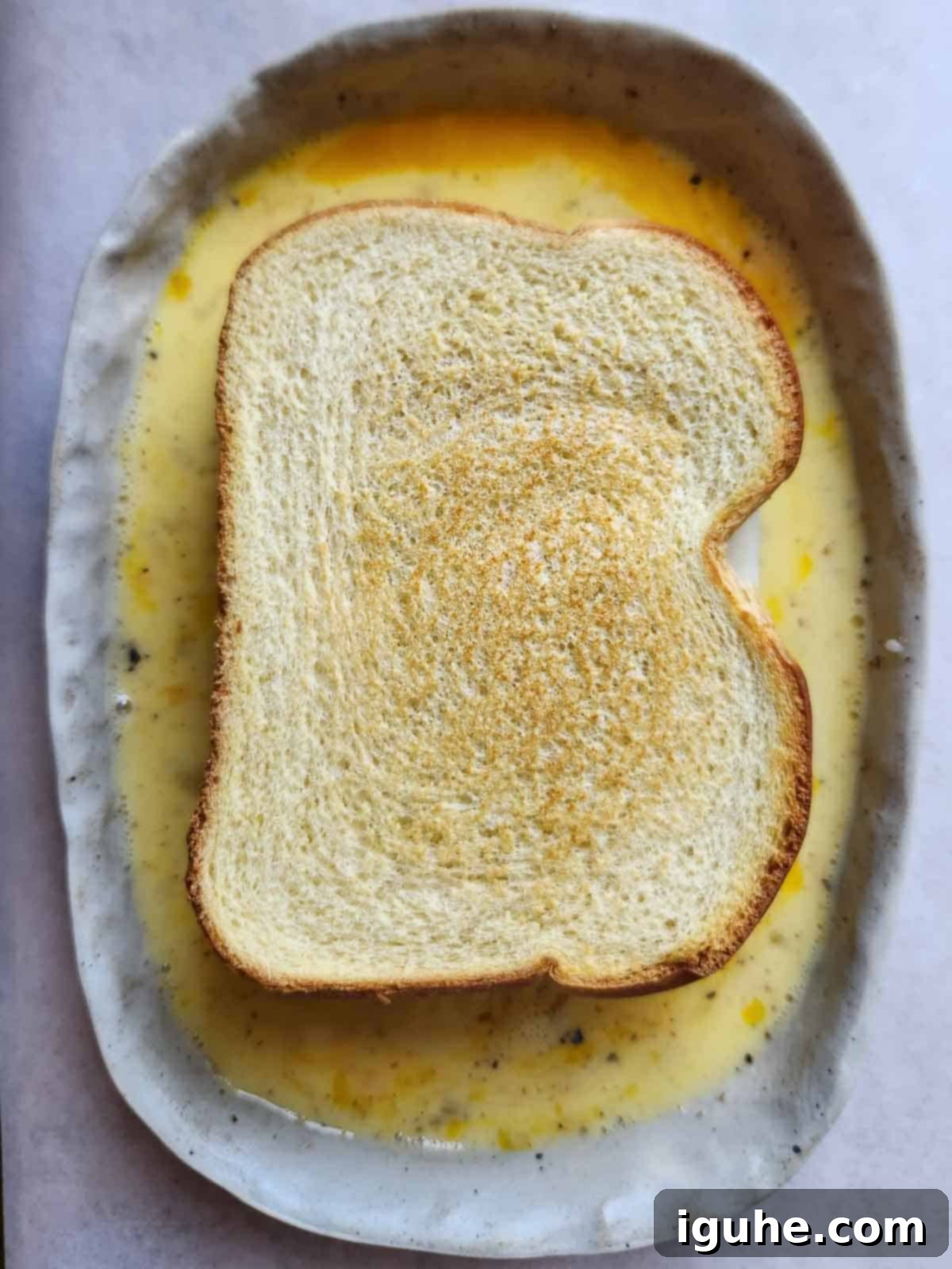 Overhead shot of a sandwich being dipped into egg custard in a shallow dish.